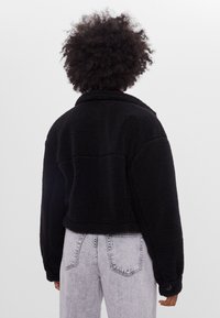 Czarna, krótka kurtka z polaru o miękkiej fakturze, z szerokimi rękawami i kołnierzem. Posiada przeszycia na plecach oraz guziki na mankietach.