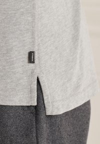 Detailaufnahme eines hellgrauen Stricktops mit kleinem schwarzem Markenetikett, getragen über dunkelgrauen strukturierten Hosen.