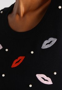 Czarny sweter z haftowanymi ustami w kolorach czerwonym, różowym i szarym, ozdobiony białymi perełkami wokół wzoru.