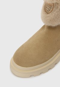 Bottine en suède beige avec une bordure en fourrure texturée, ornée d'un emblème en cristal sur le côté et dotée d'une semelle robuste.