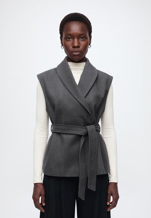 Donna che indossa un gilet grigio senza maniche con cintura sopra una maglia bianca a maniche lunghe e pantaloni neri, in piedi davanti a uno sfondo bianco uniforme.