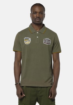 Homme portant un polo vert olive avec des écussons "Kustom Motor Oil" et "Von Dutch Motor Oil", un pantalon cargo assorti et des bijoux, sur un fond blanc.