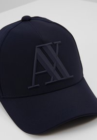 Cappellino da baseball blu navy realizzato in tessuto liscio, con un logo in rilievo "AX" dello stesso colore sul fronte e una corona arrotondata.