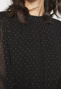 Vestido negro con mangas transparentes, que presenta una tela texturizada resaltada por pequeños puntos morados irregulares. Escote alto con pliegues.
