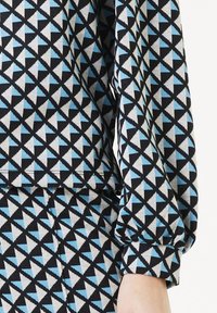 Langarm kledingstuk met een geometrisch driehoekig patroon in lichtblauw, grijs en zwart, met gathered manchetten en een gladde textuur.