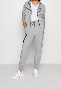 Person klädd i en grå Nike träningsoverall med zip-up huvtröja och joggers, vita sneakers och en vit T-shirt, står med händerna i fickorna.