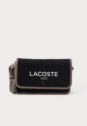 Črn tekstilni nahrbtnik čez ramo z sivimi poudarki, s pokrovom za zapiranje in belim logotipom "LACOSTE 1933" spredaj. Vključena je nastavljiva naramnica.