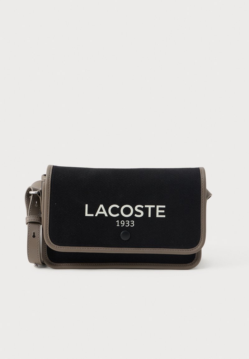 Fekete textil keresztpántos táska szürke kiegészítőkkel, amely hajtókás záródással és fehér "LACOSTE 1933" logóval rendelkezik az elején. Állítható pánt tartozik hozzá.