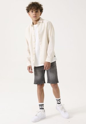 Lichtbeige overhemd over een wit T-shirt, gecombineerd met grijze denim shorts. Witte sneakers en sokken met zwarte accenten maken de outfit compleet.