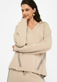 Sweatshirt beige surdimensionné avec un col en V et des manches longues. Présente des accents texturés sur les côtés et un cordon de serrage à l'ourlet.