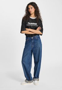 Black performance shirt with white logo, high-waisted blue denim jeans, and white sneakers with gray accents. Model has curly hair.