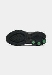 Semelle de chaussure de sport noire avec une bande de roulement texturée et trois éléments circulaires verts portant les lettres V, D et S sur un fond blanc uni.