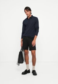 Marine langermet polo, svarte shorts, svarte sko og lyse sokker. Med en svart veske poserer modellen mot en hvit bakgrunn.