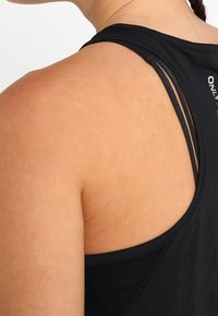 Czarny top na ramiączkach z wąskimi, skrzyżowanymi na plecach paskami oraz gładkim, elastycznym materiałem. Krój podkreśla ramiona i górną część pleców.