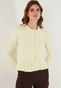 Crèmegekleurde gebreide cardigan met een ronde halslijn, knoopsluiting aan de voorkant en een gerimpelde zoom. Gemaakt van zachte stof met lange mouwen en parelknopen.