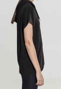 Svart huvtröja med korta ärmar, tillverkad av mjukt material, med en avslappnad passform och en rundad nederkant. Minimalistisk design utan synliga mönster.