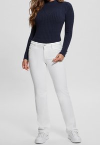 Marineblaue, gerippte Langarmbluse mit hohem Kragen, kombiniert mit weißen, gerade geschnittenen Jeans und weißen Sneakern.