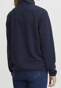 Sudadera de forro polar azul marino con cuello alto, que presenta una textura suave y un diseño ajustado. Detalle de logo pequeño en el dobladillo.