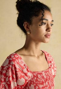 Rotes Oberteil mit Blumenmuster, quadratischem Ausschnitt, Puffärmeln und gerafftem Textur. Getragen mit goldenen Ohrringen und gestyltem Haar.