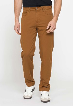 Uomo con pantaloni marroni a gamba dritta e sneaker bianche con strisce nere, in piedi davanti a uno sfondo chiaro e uniforme.