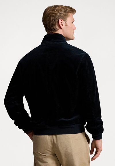 Giacca in corduroy nera con colletto a camicia, polsini e orlo a costine, che presenta una superficie testurizzata. Indossata sopra pantaloni beige.