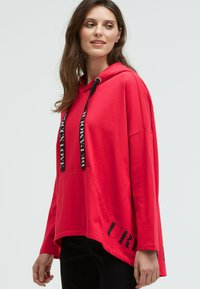 Roter Kapuzenpullover mit lockerer Passform, ausgestattet mit schwarzen Kordeln und Schriftzügen. Weicher Stoff, Kängurutasche und längerer Saum hinten.