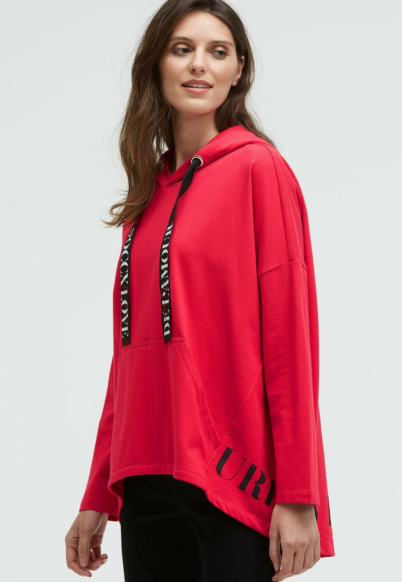 Roter Kapuzenpullover mit lockerer Passform, ausgestattet mit schwarzen Kordeln und Schriftzügen. Weicher Stoff, Kängurutasche und längerer Saum hinten.