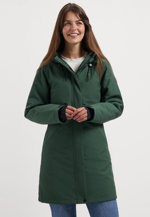 Frau mit langen braunen Haaren, die einen dunkelgrünen winterlichen Kapuzenmantel und blaue Jeans trägt, lächelt und blickt leicht zur Seite.