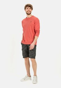 Koraalkleurige sweatshirt met lange mouwen en logo, gecombineerd met zwarte shorts met een bladerenpatroon, en witte sneakers.