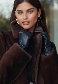 Barna szőrme bundával, széles gallérral, fekete bőrkesztyűvel és arany geometrikus fülbevalókkal. Simára csiszolt textúra és meleg dizájn részletek láthatók.