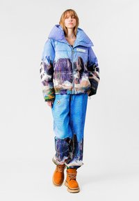 Chaqueta acolchada con estampado de paisaje montañoso en tonos azules y púrpuras, combinada con pantalones texturizados azules y botas marrones con cordones.