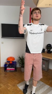 Biely a čierny športový dres s nápisom "Highperformance", ružové šortky, žonglujúci s kartónovou krabicou vo vnútri. Viditeľná drevená podlaha a rastliny.