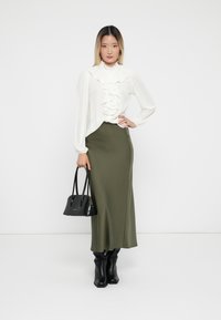 White blouse with ruffled detailing, olive green skirt, black knee-high boots, and a small black handbag. Smooth textures and fitted silhouette.