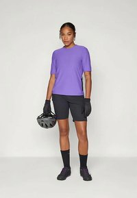 Mulher vestindo uma camisa roxa de manga curta, calções de ciclismo pretos, luvas, meias e sapatos, segurando um capacete de bicicleta preto ao seu lado.