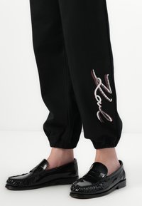 Sorte joggebukser med elastiske mansjetter har dekorativt broderte tekst. Glossy sorte loafers med strukturert frynser og flat såle.