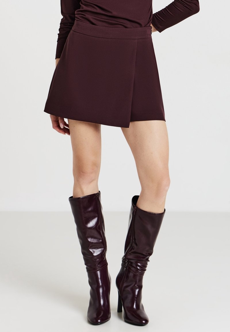 Femme portant une jupe portefeuille bordeaux foncé et des bottes hautes en cuir assorties à talons, debout avec une main sur la hanche.