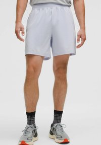 Un homme portant un short de sport gris clair, des chaussettes à motif noir et gris, et des chaussures de course grises avec des semelles orange, se tenant contre un fond uni.