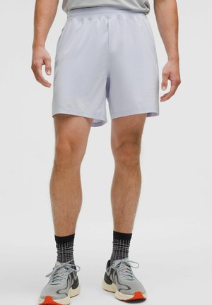 Mann in hellgrauen Sportshorts, schwarzen und grauen gemusterten Socken und grauen Sportschuhen mit Orangeböden, der vor einem einfarbigen Hintergrund steht.