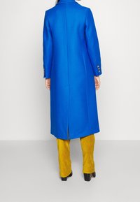 Cappotto lungo in lana blu reale con silhouette aderente, colletto a punta e polsini abbottonati, abbinato a pantaloni in corduroy color senape.