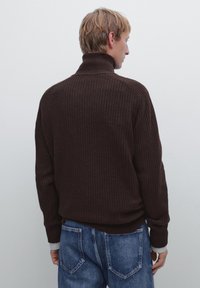 Brązowy sweter typu golf z prążkowaną strukturą, o luźnym kroju i wysokim kołnierzu, zestawiony z niebieskimi dżinsami. Miękka tekstura z kontrastującymi mankietami.