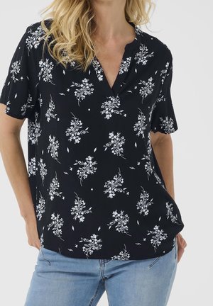 Vrouw draagt een zwarte blouse met korte mouwen en witte bloemenprint en lichtblauwe spijkerbroek, staand met handen in de zakken.