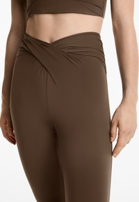Bruna höga leggings tillverkade av stretchigt tyg med en vriden framsida och slät yta. Inga synliga mönster eller metalldetaljer.