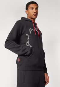 Svart hoodie med röda dragsnören, med vit grafisk design framtill och ett röd emblem på den nedre fickområdet. Mjuk tyg.