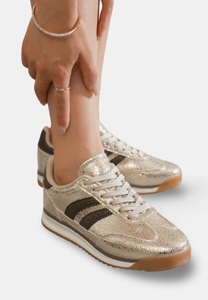 Baskets dorées métalliques avec une finition texturée, des rayures brunes et une semelle blanche. Elles sont dotées de lacets et d'un col rembourré pour le confort.
