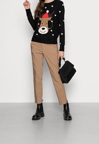 Pull noir avec motif de renne et pois blancs, pantalon beige sur mesure, bottines Chelsea noires, et sac à main noir avec une poignée tressée.