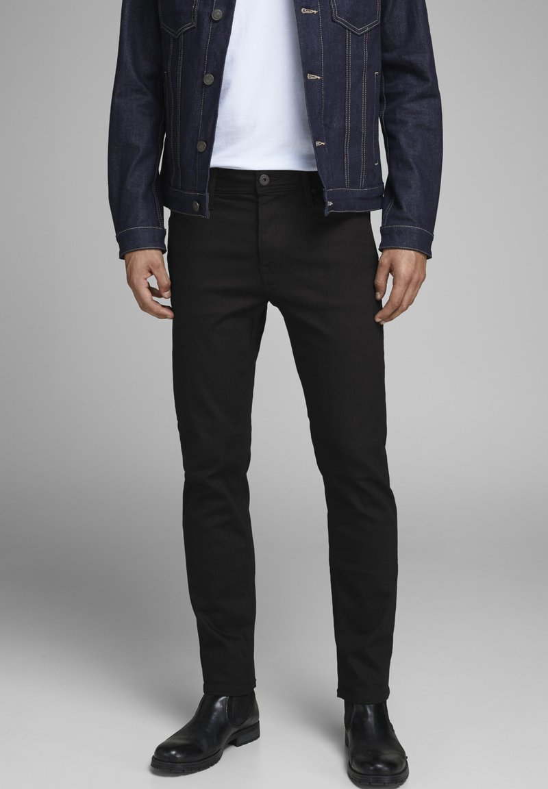 Mustad slim-fit teksad, millel on sile tekstuur, koos valge t-särgi ja tume teksajaki ning mustade hõlmadega saapatega. Hõlmadega saapad täiendavad riietust.