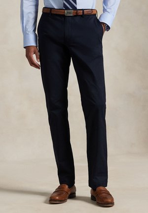 Mann trägt marineblaue Anzughose, hellblaues Hemd mit gestreifter Krawatte, braunen Ledergürtel, Armbanduhr und braune Lederloafers.