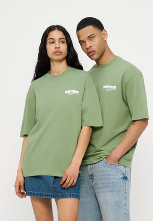 Dvaja modely nosia zladené zelené oversize tričká s logom „MATCHA“, žena v rifľovej sukni, muž v svetlomodrých džínsoch, neutrálne výrazy tváre.