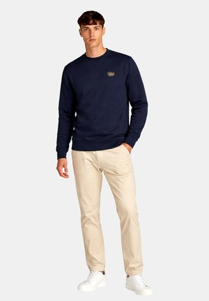 Ung mand iført en marineblå sweatshirt med lille logo, beige bukser og hvide sneakers, stående med den ene hånd i lommen.