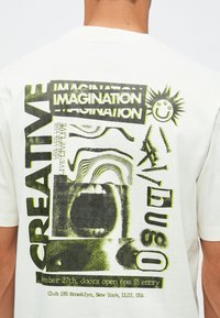 Baksidan av en vit t-shirt med svart och gul grafisk design som visar en stor mun, "KREATIV," "FANTASI," och detaljer om evenemanget för Club 185 Brooklyn.